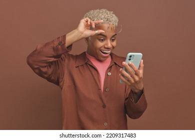 Young emotional cheerful African American woman lifts glasses from eyes and opening mouth looks at screen of mobile phone when sees long-awaited message from friend or buddy stands in brown studio - Shutterstock ID 2209317483