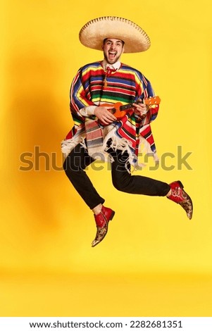 Young emotional man in colorful poncho and sombrero posing, playing guitar and jumping against yellow studio background. Concept of mexican traditions, fun, celebration, festival, emotions