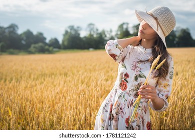 young, emotional, pretty girl in a light dress with a light hat at sunset in a wheat, golden field, holding ears of wheat 