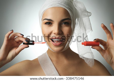 Young emotional woman with a gauze bandage on her head and chest, holding lip gloss, on grey background