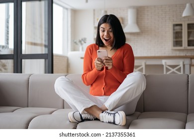 Young Emotional Woman Holding Smartphone Playing Mobile Game Celebration Success At Home. Excited African American Hipster Using Mobile Phone Shopping Online With Big Sales Sitting On Sofa