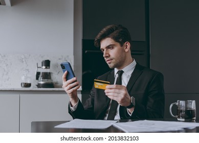 Young employee business man corporate lawyer in black suit shirt tie read paper document using mobile cell phone credit bank card shopping online order delivery booking tour sit at table in kitchen.