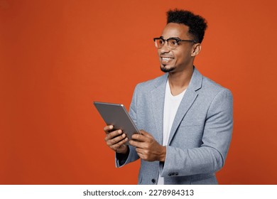 Young employee business man corporate lawyer wears classic formal grey suit shirt glasses work in office use digital tablet pc computer look aside area isolated on plain red orange background studio