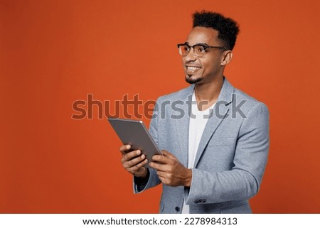 Young employee business man corporate lawyer wears classic formal grey suit shirt glasses work in office use digital tablet pc computer look aside area isolated on plain red orange background studio