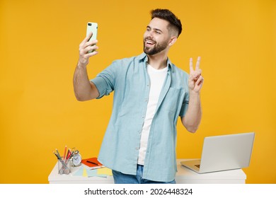 Young employee business man in shirt stand work at white office desk pc laptop do selfie shot on mobile phone post photo on social network show victory gesture isolated on yellow background studio.