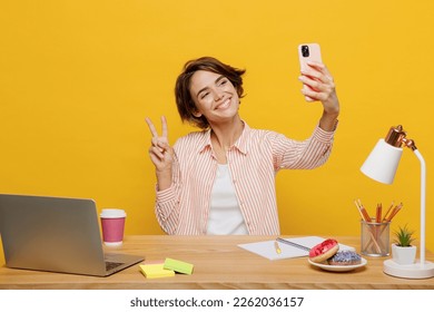 Young employee business woman wear casual shirt sit work at office desk with pc laptop do selfie shot mobile cell phone show v-sign isolated on plain yellow color background Achievement career concept