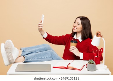 Young employee business woman wear red sweater sit work at office put legs on desk with pc laptop do selfie shot on mobile cell phone drink coffee isolated on plain beige background. Career concept