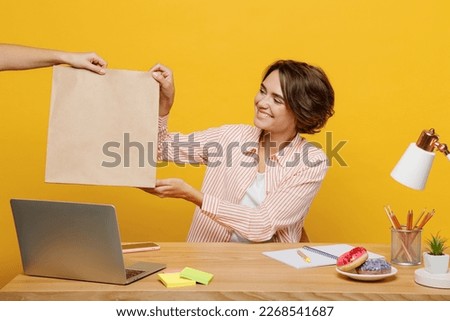 Young employee business woman wear casual shirt sit work at office desk with pc laptop receive take brown clear blank craft paper takeaway bag isolated on plain yellow color background. Career concept