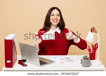 Young employee business woman wear red sweater sit work at office desk with pc laptop hold store gift certificate coupon voucher card show thumb up isolated on plain beige background. Career concept