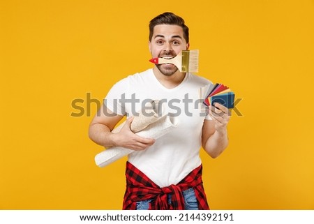 Young employee handyman man wear t-shirt hold color palette paint wallpaper rolls biting brush isolated on yellow background. Instruments accessories renovation apartment room. Repair home concept.