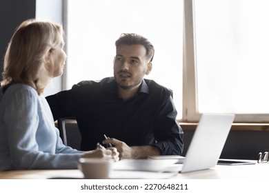 Young employee man, intern consulting senior mentor woman. Two diverse colleagues sitting at work table, talking at laptop, discussing project plan, strategy, task, teamwork - Powered by Shutterstock