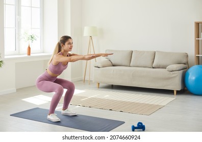 Young Enduring Woman Trains At Home Doing Deep Squats With Her Arms Outstretched. Slender Woman In Comfortable Pink Sportswear Does Morning Workout Standing On Sports Mat In Living Room. Sport Concept