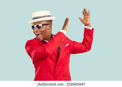 Young energetic African American man wearing sunglasses and red entertainment suit, yelling and clapping hands performing funny dance or working as host of party stands on pastel blue background