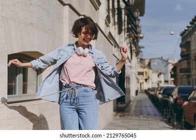 Young energetic fun excited caucasian woman 20s in jeans clothes headphones eyeglasses listening to music dancing outdoors on city street sidewalk walk stroll. People urban youth lifestyle concept.