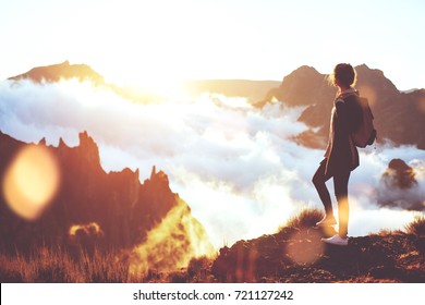Young energetic girl with  backpack standing at edge of cliff in mountains and looking at sunset with a clouds.