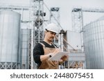 A young engineer in a hard hat with a project in his hands checks the status of the silo construction.