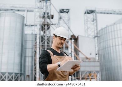 A young engineer in a hard hat with a project in his hands checks the status of the silo construction.