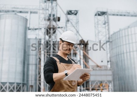 A young engineer in a hard hat with a project in his hands checks the status of the silo construction.