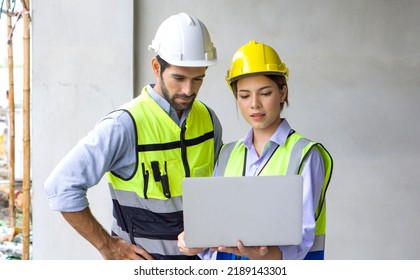Young Engineer With Laptop Computer Explain To Foreman About A Floor Plan. Both Wear Construction Helmet And Safety Vest. Work Environment At The Construction Site Of Housing Projects.