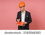 a young engineer in a protective helmet and jacket points inquiringly at a notebook in his hands on a pink background