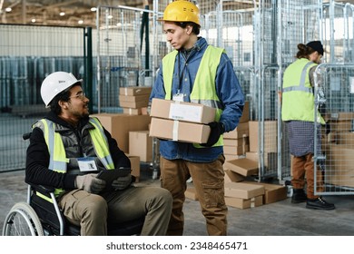 Young engineer with stack of packed cardboard boxes talking to colleague sitting in wheelchair and using tablet during discussion of working points - Powered by Shutterstock