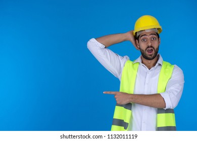 Young engineer standing hand behind head pointing with one finger shocked on a blue background.