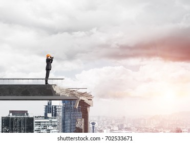 Young engineer in suit and helmet looking in binoculars while standing on broken bridge with cityscape and sunlight on background. 3D rendering.