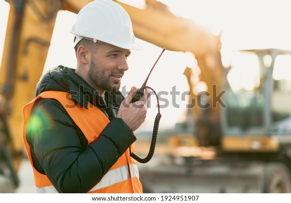 Young\
engineer talking radio communication (walkie talkie) and wearing a\
white helmet and construction orange vest. Close up engineers\
working on a building site with sunny\
background.