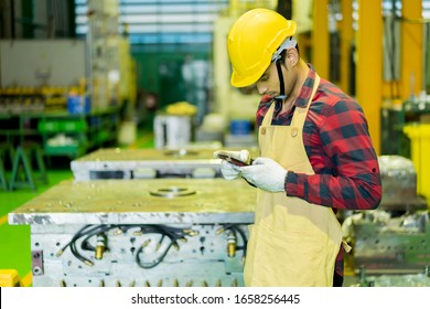 Young Engineer In Uniform With Safety Helmet Looking At Smartphone While Upload Data Or Sending Text Order Confirm To Customer. Worker Uses Mobile Phone While Relax On Industry Manufacturing Factory 