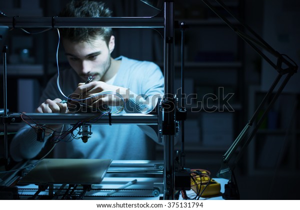 Young engineer working at night in the lab, he is\
adjusting a 3D printer\'s components, technology and engineering\
concept