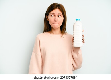 Young English woman holding a bottle of milk isolated on blue background confused, feels doubtful and unsure.