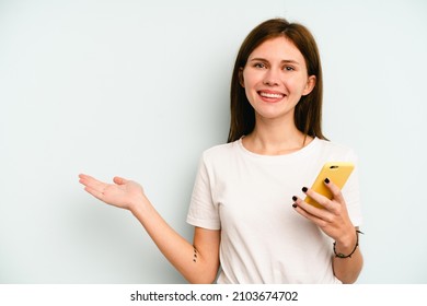 Young English Woman Holding Mobile Phone Isolated On Blue Background Showing A Copy Space On A Palm And Holding Another Hand On Waist.