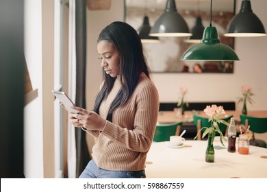 Young Entrepreneur Is Reading E-mail On The Screen Of The Digital Tablet While Standing Beside The Window In A Cafe With Modern Interior. African Woman Is Surfing The Web On A Portable Computer.