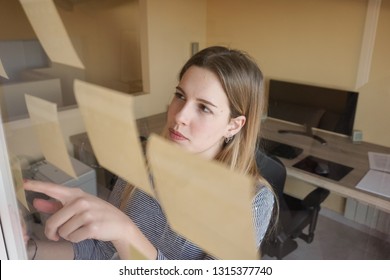 Young entrepreneur woman is brainstorming ideas on sticky notes on the window or glass wall. It is the Agile methodology for writing stories with the story mapping meeting of her product backlog. - Shutterstock ID 1315377740
