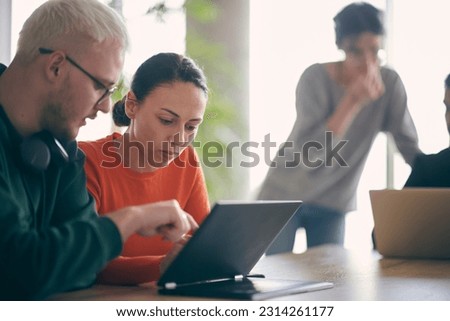 A young entrepreneurial couple sits together in a large, modern office, engaged in analyzing statistics and data on their laptop