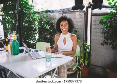 Young ethnic female in casual clothes sitting at table with laptop smartphone and notebook looking at camera and laughing brightly - Powered by Shutterstock