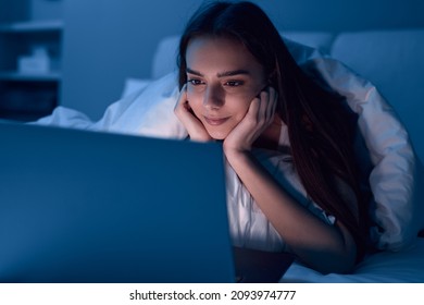 Young ethnic female with long dark hair lying on bed with hands on cheeks while watching interesting movie on laptop at night at home