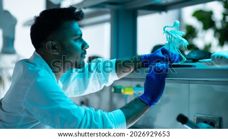 Young Ethnic Guy Working in a Modern Laboratory Wearing a White Lab Coat and Blue Surgical Gloves. Lab Assistant Looking at an Animal Skeleton, Examining and Inspecting it.