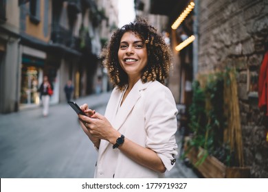 Young ethnic woman with smartphone smiling and standing on street and browsing internet wearing casual clothes on sunny day looking forward