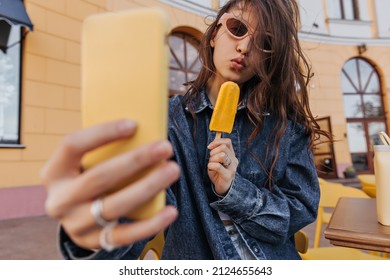 Young european brown-haired girl makes selfie on modern smartphone outdoors. Cute teenager in sunglasses holds ice cream, dressed in denim jacket. Cell phone use concept