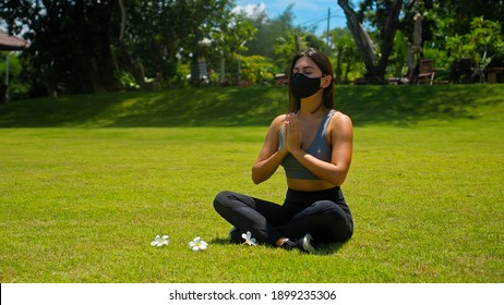 Young European brunette girl practices alone yoga in nature sitting on green grass in lotus pose wearing a black protective mask on her face - Powered by Shutterstock