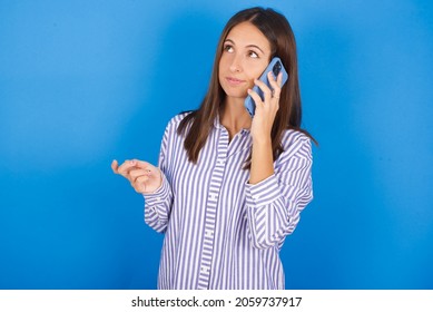 Young European Brunette Woman Wearing Stripped Shirt On Blue Background Speaks On Mobile Phone Spends Free Time Indoors Calls To Friend.