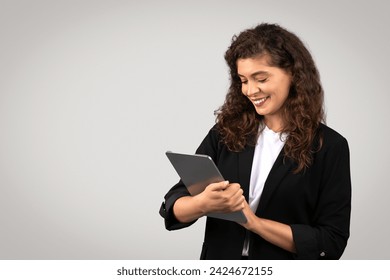 Young european businesswoman in black suit using digital tablet, reviewing important documents or chatting online with clients, posing on gray studio background, free space - Powered by Shutterstock