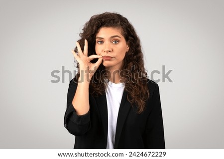 Young european businesswoman in black suit making silence gesture with her finger on her lips, signaling the need for quiet or secrecy, looking at camera on grey backdrop