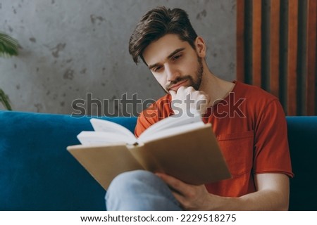 Young european calm man 20s wears red t-shirt read book novel sit on blue sofa couch stay at home hotel flat rest relax spend free spare time in living room indoors grey wall. People lounge concept