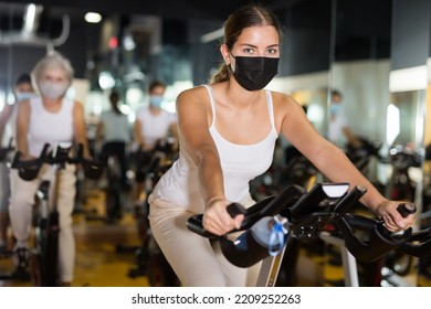 Young European Female In Protective Mask Riding Exercise Bike During Cycling Class In Gym