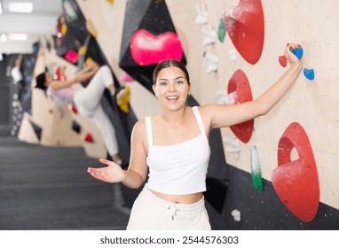 Young European girl poses near steep climbing wall in gym and leans against hook. Woman is resting after workout and leaning against artificial wall for sports activities - Powered by Shutterstock