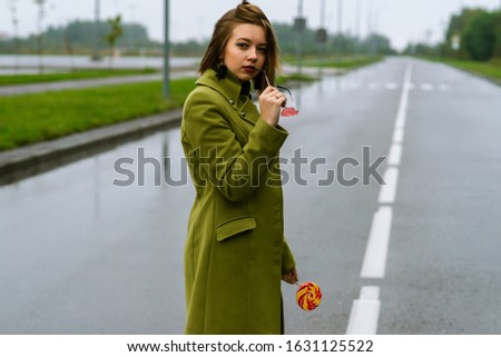 young european girl walks in rainy weather down the street with puddles of green cloak and eats lick