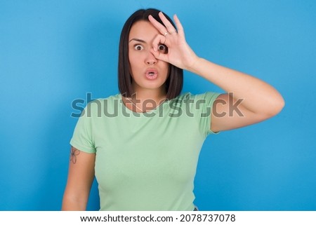 Young european girl wearing green T-shirt over blue background doing ok gesture shocked with surprised face, eye looking through fingers. Unbelieving expression.