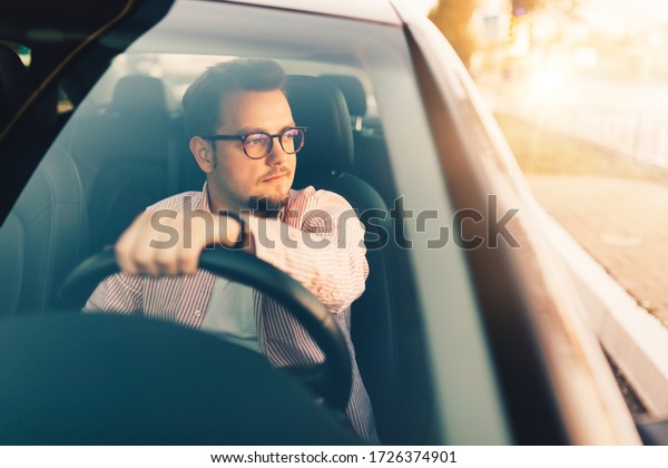 A young\
european happy driver in comfortable \
expensive car. Stylish man\
wearing glasses. View through the\
windshield